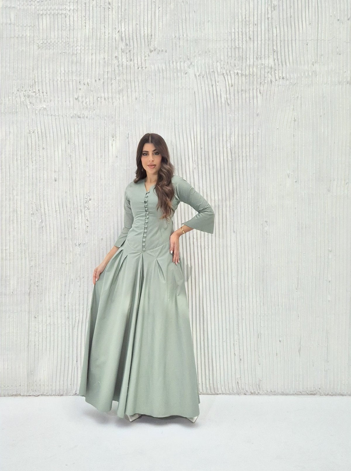 Model wearing a sage green pleated maxi dress with a buttoned bodice in a minimalist background